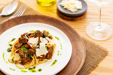 Beef ragout sauce spaghetti pasta with tomatoes, basil and pesto. Aside cutlery, a bottle of olive oil, parmesan and wine glass. White plate on a wooden plate on a jute mat on an oak table.