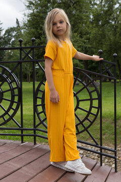 A little girl of six-seven years old with long blond hair in a plain yellow outfit stands and smiles in a summer park on a decorative bridge and poses in full growth.