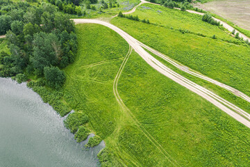 countryside landscape in summer. rural dirt road on green meadow near lake. birds eyes view