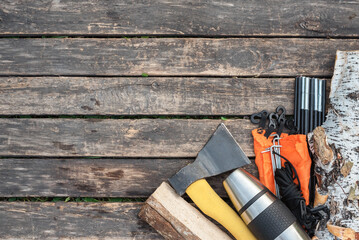 Stainless cup, parts of tent, vacuum flask, axe, birch logs and rope on the wooden table background with copy space. Camping.