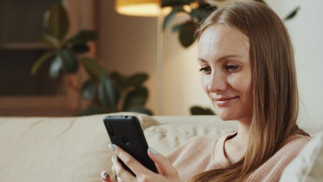 Tilt up shot of beautiful blonde woman sitting on couch at home, swiping on smartphone screen and smiling while checking social media