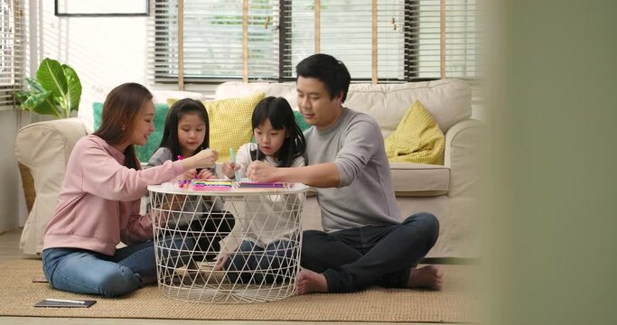 Happy young asian family parents and cute little children drawing coloring picture with pencils helping cute child daughter enjoying talk play at home, mom dad and kid girl having fun in living room