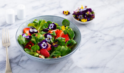Fresh green salad with strawberries and edible flowers in a bowl. Marble background. Copy space.