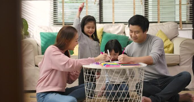 Happy young asian family parents and cute little children drawing coloring picture with pencils helping cute child daughter enjoying talk play at home, mom dad and kid girl having fun in living room