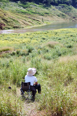 Alone Asian Thai Woman relaxing and sitting on camping chair in the grass field near Stream in tropical forest of Thailand. Lifestyle and travel Concep