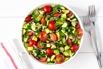 Salad of fresh ripe tomato, onion, avocado, cucumber and parsley and olive oil, salad in a white plate with a fork and towel on a white wooden background