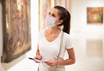 Focused adult girl in disposable face mask admiring paintings in museum holding brochure with...