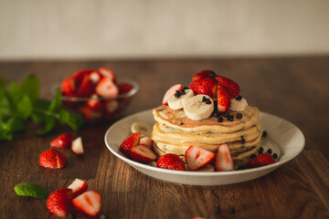 stacked pancakes on a plate looks very delicious. the pancakes have bananas and strawberries on them and look very sweet. there is also syrup pouring down on the pancakes and looks very healthy. 
