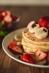 stacked pancakes on a plate looks very delicious. the pancakes have bananas and strawberries on them and look very sweet. there is also syrup pouring down on the pancakes and looks very healthy. 
