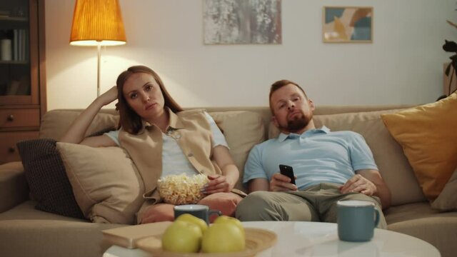 Bored Married Couple Resting Together On Couch In The Living Room, Eating Popcorn And Switching Channels With Remote Control While Watching TV At Home