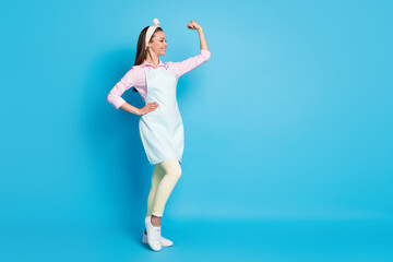 Full length photo of attractive lady confident house wife good mood showing strong muscle arm fist wear headband long dotted apron pink shirt pants shoes isolated blue color background