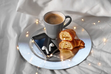 morning, hygge and breakfast concept - smartphone, wireless earphones, cup of coffee and croissants with garland lights on plate in bed at home