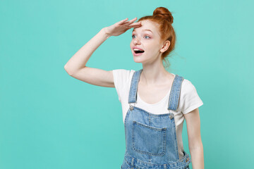 Amazed young readhead girl in casual denim clothes posing isolated on blue turquoise background studio. People lifestyle concept. Mock up copy space. Hold hand at forehead looking far away distance.