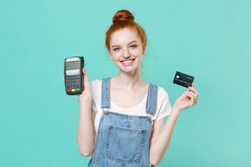 Funny young readhead girl in casual denim clothes isolated on blue turquoise background. People lifestyle concept. Hold wireless modern bank payment terminal to process acquire credit card payments.