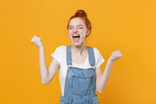 Happy young readhead girl in casual denim clothes white t-shirt isolated on yellow background studio portrait. People lifestyle concept. Mock up copy space. Doing winner gesture, keeping eyes closed.
