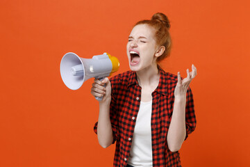 Crazy young readhead girl in casual red checkered shirt posing isolated on orange background. People lifestyle concept. Mock up copy space. Screaming on megaphone keeping eyes closed, spreading hands.