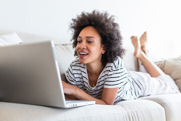 Young beautiful woman with curly hair working on laptop computer while sitting on the sofa at home, heck on oline shops for cyber monday sales. Technology woman concept alternative office freelance