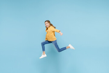 Full length portrait of amazed little blonde kid girl 12-13 years old in yellow jacket isolated on pastel blue background studio. Childhood lifestyle concept. Mock up copy space. Jumping like running.