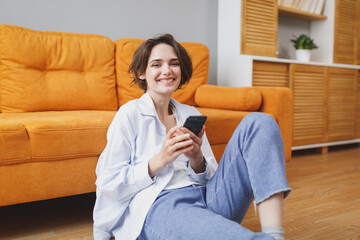 Beautiful young woman girl in white casual clothes sit on floor near couch spending time in living room at home. Rest relax good mood leisure lifestyle concept. Using mobile phone, typing sms message.