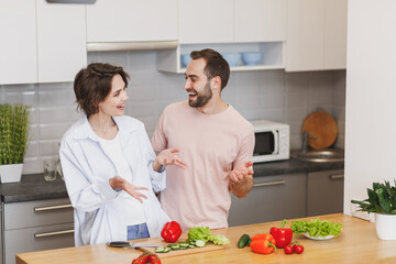 Laughing young couple friends guy girl in casual clothes preparing vegetable salad cooking food in kitchen at home. Dieting family healthy lifestyle concept. Mock up copy space. Speaking talking.