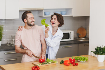 Laughing funny young couple two friends guy girl in casual clothes preparing vegetable salad cooking food in light kitchen at home. Dieting family healthy lifestyle concept. Mock up copy space.