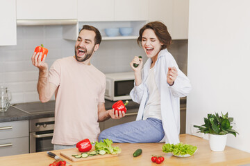 Laughing couple friends guy girl sit on table preparing vegetable salad cooking food in light kitchen at home. Dieting family healthy lifestyle concept. Mock up copy space. Hold cucumber bell peppers.