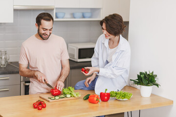 Funny young couple two friends guy girl in casual clothes sit on table preparing vegetable salad cooking food in light kitchen at home. Dieting family healthy lifestyle concept. Mock up copy space.