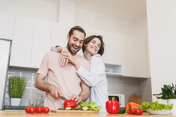 Smiling young couple two friends guy girl in casual clothes preparing vegetable salad cooking food in light kitchen at home. Dieting family healthy lifestyle concept. Mock up copy space. Hugging.