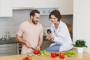 Excited young couple friends guy girl sit on table preparing vegetable salad cooking food in light kitchen at home. Dieting family healthy lifestyle concept. Mock up copy space. Using mobile phone.