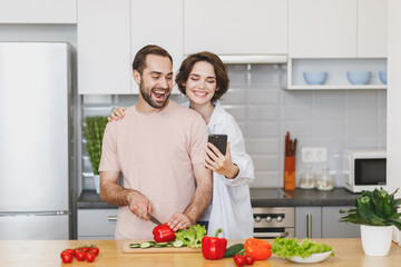 Funny young couple friends guy girl preparing vegetable salad cooking food in light kitchen at home. Dieting family healthy lifestyle concept. Mock up copy space. Doing selfie shot on mobile phone.
