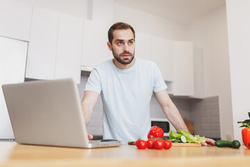 Attractive young man in casual t-shirt using laptop computer looking at recipe preparing vegetable salad cooking food in light kitchen at home. Dieting healthy lifestyle concept. Mock up copy space.
