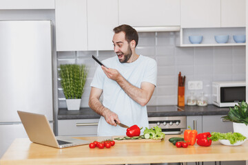 Excited young bearded man guy in white casual t-shirt talking on mobile phone preparing vegetable salad cooking food in light kitchen at home. Dieting healthy lifestyle concept. Mock up copy space.