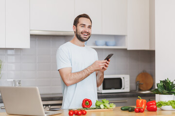 Funny young bearded man in white t-shirt using mobile phone looking for recipe preparing vegetable salad cooking food in light kitchen at home. Dieting healthy lifestyle concept. Mock up copy space.