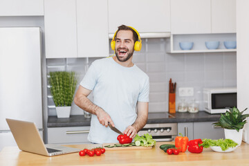 Cheerful young bearded man in white casual t-shirt listen music with headphones preparing vegetable salad cooking food in light kitchen at home. Dieting healthy lifestyle concept. Mock up copy space.