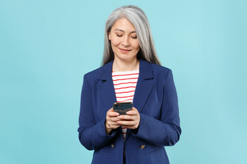 Smiling gray-haired business woman in blue suit posing isolated on pastel blue background studio. Achievement career wealth business concept. Mock up copy space. Using mobile phone typing sms message.
