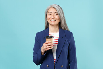 Smiling gray-haired business woman in blue suit posing isolated on blue background studio portrait. Achievement career wealth business concept. Mock up copy space. Hold paper cup of coffee or tea.