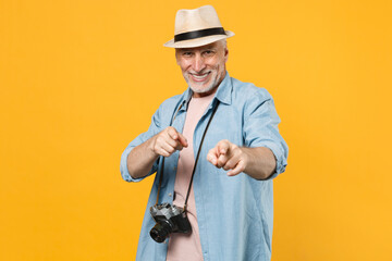 Smiling traveler tourist elderly gray-haired man in hat photo camera isolated on yellow background. Passenger traveling abroad on weekend. Air flight journey concept. Pointing index fingers on camera.