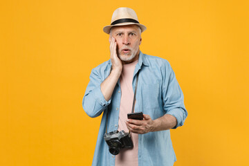 Amazed traveler tourist elderly gray-haired man isolated on yellow background. Passenger traveling abroad on weekends. Air flight journey. Using mobile cell phone booking taxi hotel put hand on cheek.