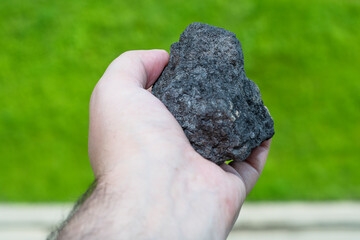 Male hand with a piece of coal on a blurred background of green forest. An image on the theme of geology and the extraction of minerals and natural fuels.