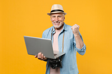 Happy traveler tourist elderly gray-haired man isolated on yellow background. Passenger traveling abroad on weekend. Air flight journey Work on laptop computer booking taxi hotel doing winner gesture.