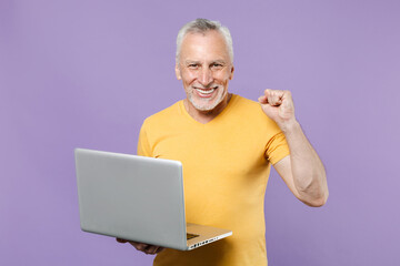 Joyful elderly gray-haired mustache bearded man in yellow t-shirt isolated on violet wall background. People lifestyle concept. Mock up copy space. Working on laptop pc computer doing winner gesture.