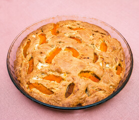 A large, round baked apple and apricot pie stands in a glass form on a table with a tablecloth.