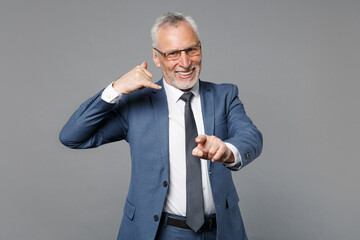 Smiling elderly gray-haired business man in blue suit isolated on grey background. Achievement career wealth business concept. Doing phone gesture says call me back, pointing index finger on camera.