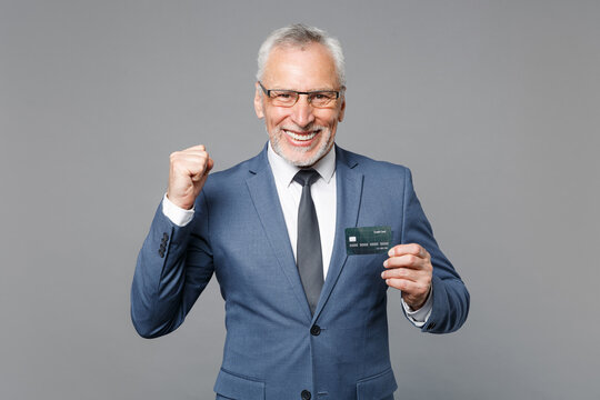 Happy elderly gray-haired business man in classic blue suit shirt tie isolated on grey wall background studio. Achievement career wealth business concept. Hold credit bank card, doing winner gesture.