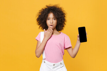 Shocked african american kid girl 12-13 years old in pink t-shirt isolated on yellow background. Childhood concept. Mock up copy space. Hold mobile phone with empty screen, covering mouth with hand.