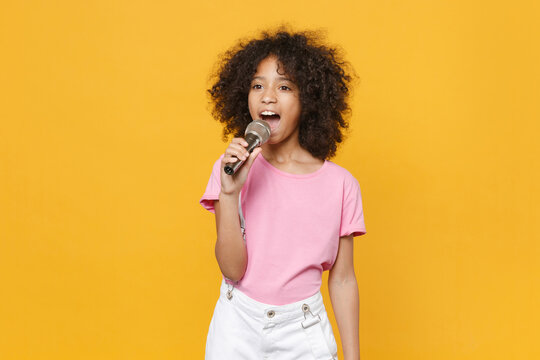 Charming little african american kid girl 12-13 years old in pink t-shirt isolated on yellow wall background studio portrait. Childhood lifestyle concept. Mock up copy space. Sing song in microphone.