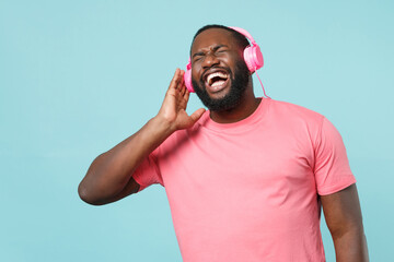 Naklejka premium Cheerful young african american man in casual pink t-shirt isolated on blue background studio portrait. People lifestyle concept. Mock up copy space. Listen music with headphones keeping eyes closed.