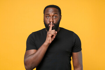 Secret african american man guy football fan in casual black t-shirt isolated on yellow background. People lifestyle concept. Mock up copy space. Saying hush be quiet with finger on lips shhh gesture.