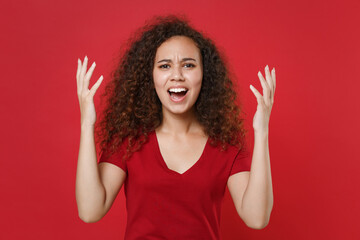 Angry young african american woman girl in casual t-shirt posing isolated on red background studio portrait. People emotions lifestyle concept. Mock up copy space. Spreading hands, screaming swearing.