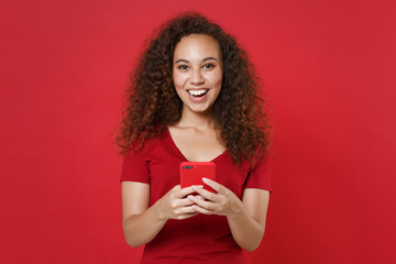 Cheerful young african american girl in casual t-shirt posing isolated on red wall background studio portrait. People lifestyle concept. Mock up copy space. Using mobile cell phone typing sms message.